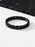A black beaded stretch bracelet made of glossy, rectangular faceted beads, displayed on a clean white surface with a dark black marble slate prop in the background.