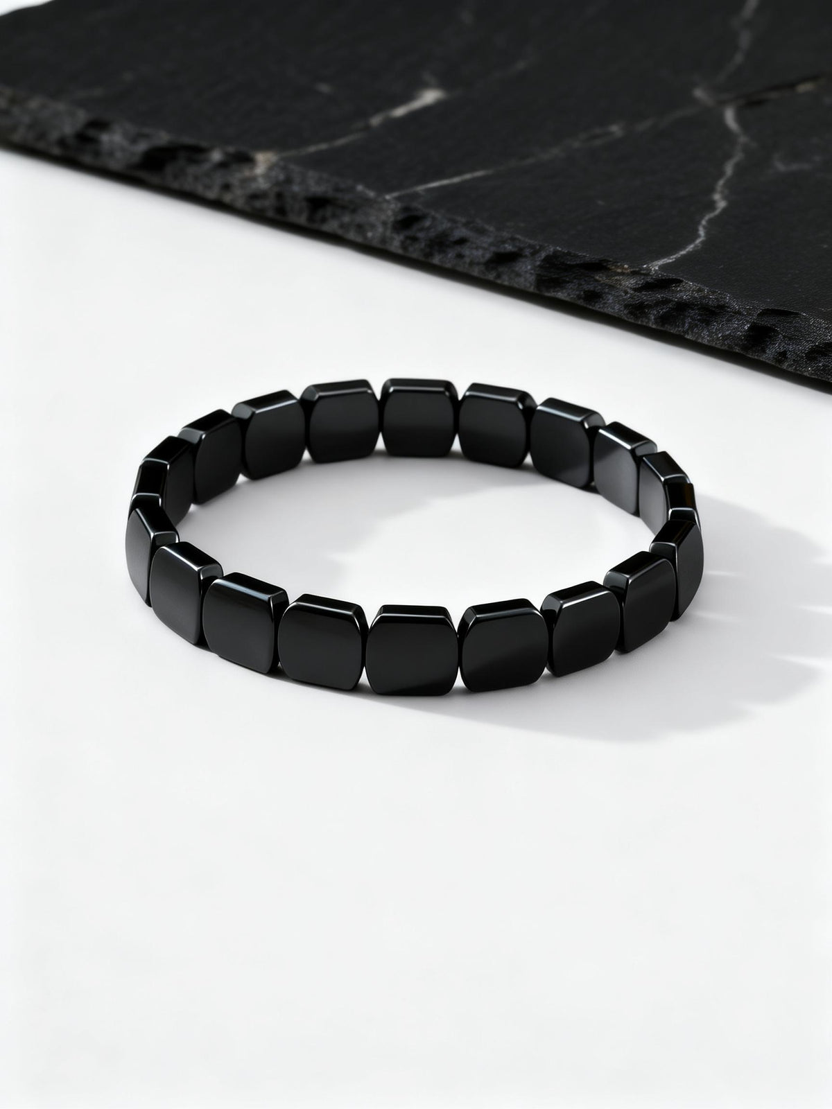 A black beaded stretch bracelet made of glossy, rectangular faceted beads, displayed on a clean white surface with a dark black marble slate prop in the background.