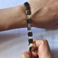 A close-up of a bracelet worn on a wrist, with fingers holding the lower end of the bracelet to show its flexibility. The bracelet features glossy, dark metallic square beads on a thin silver-toned band.