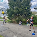 Three children playing with tennis balls and tennis rackets on a paved backyard court in front of a large evergreen tree.