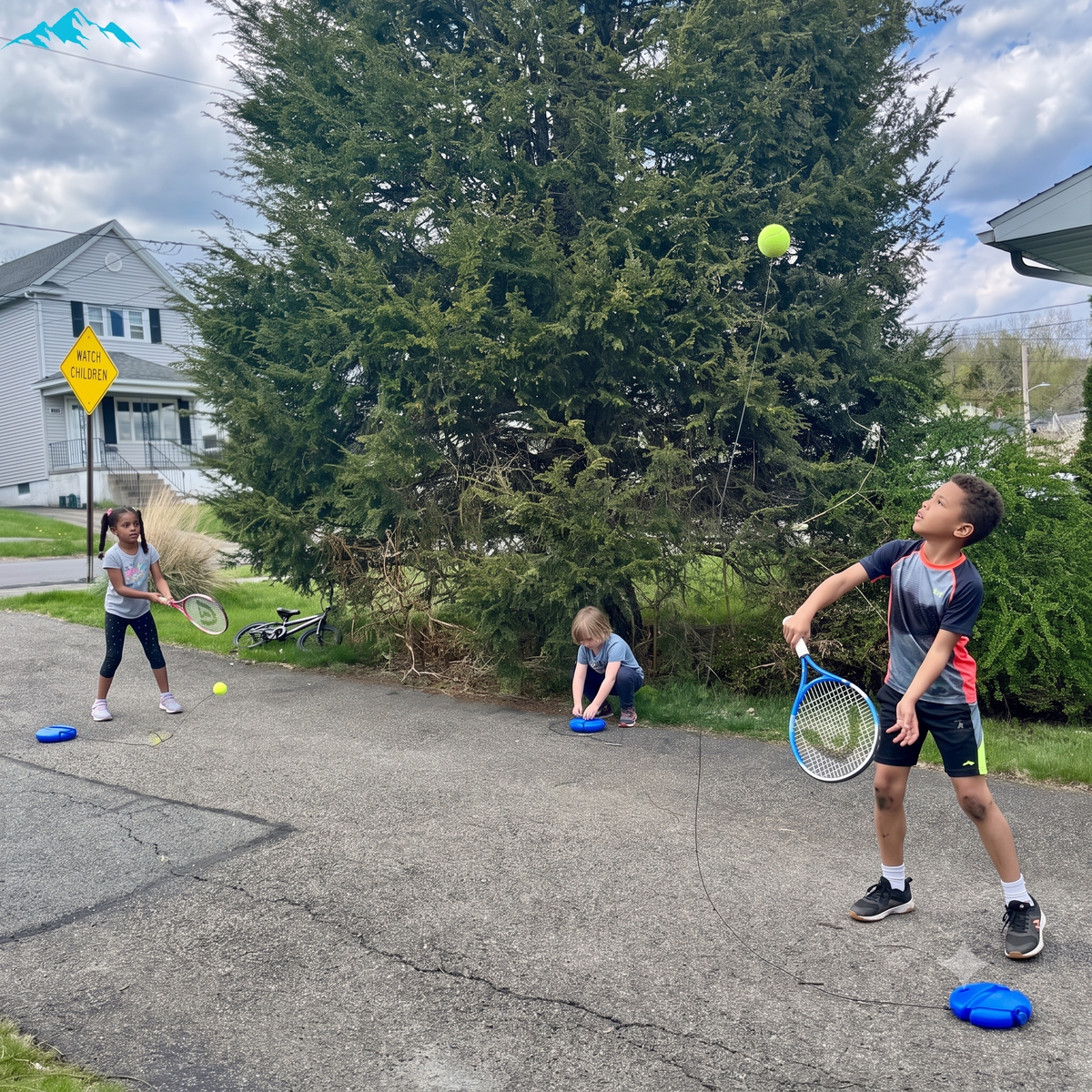 Three children playing with tennis balls and tennis rackets on a paved backyard court in front of a large evergreen tree.
