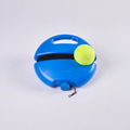 A blue tennis ball retriever tool with a handle, black elastic band, and a yellow tennis ball resting on top, photographed on a light gray studio background.